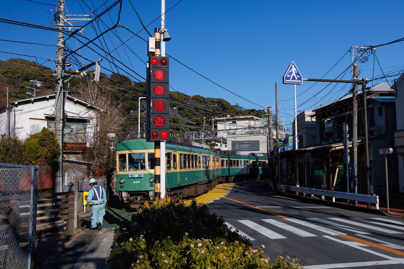 江ノ電ニキの聖地です。踏切の手前に「電車接近」と赤色で点灯した警報機があります。奥には緑と黄色で塗装された江ノ電がカーブを通過しています。