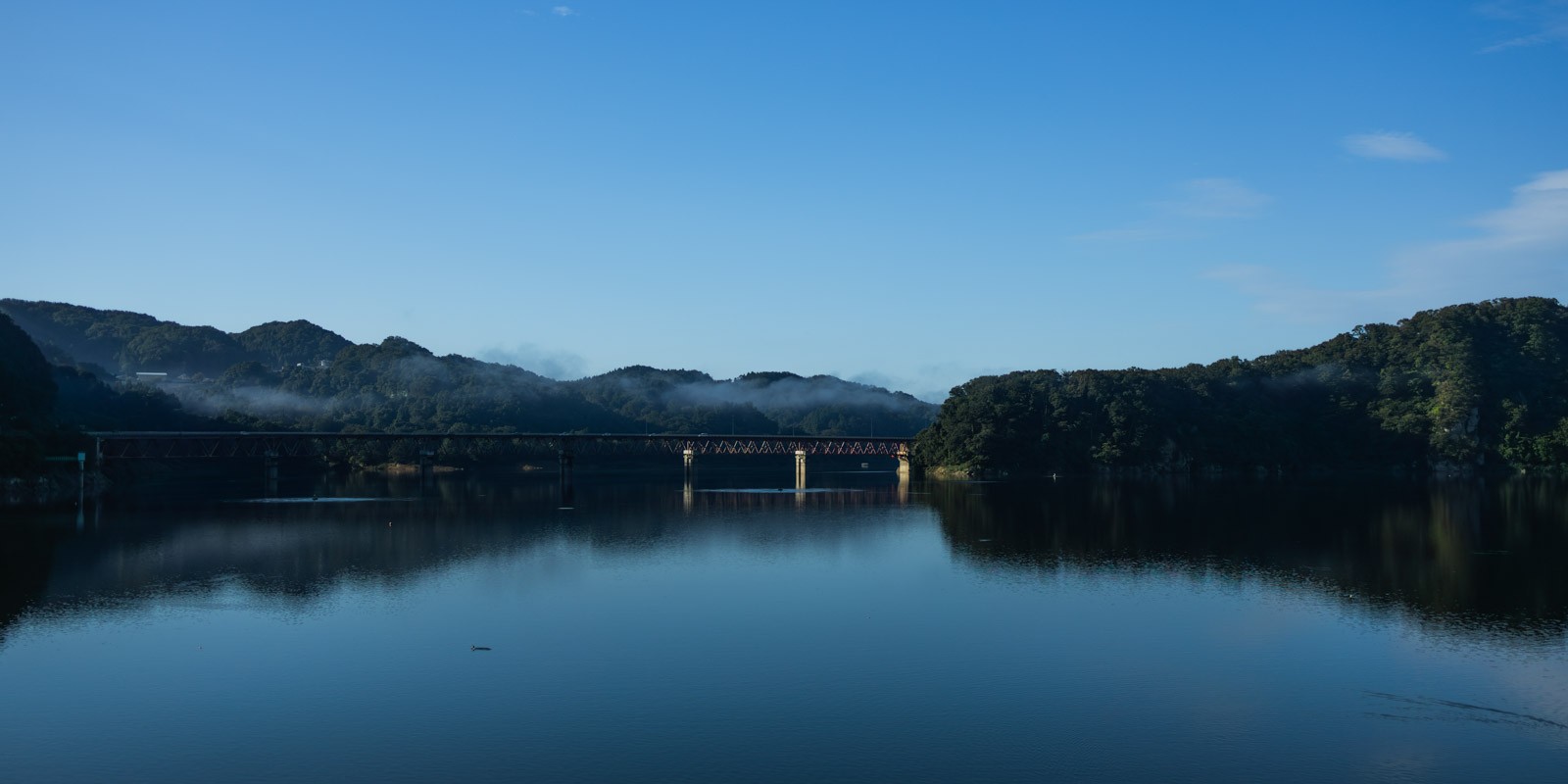 釜房ダムに釜房大橋が架かり、その姿が水面に映り込んでいます。橋の向こうには蔵王の山々がそびえています。空は澄んだ青色をしています。
