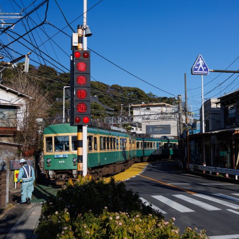 江ノ電ニキの聖地です。踏切の手前に「電車接近」と赤色で点灯した警報機があります。奥には緑と黄色で塗装された江ノ電がカーブを通過しています。