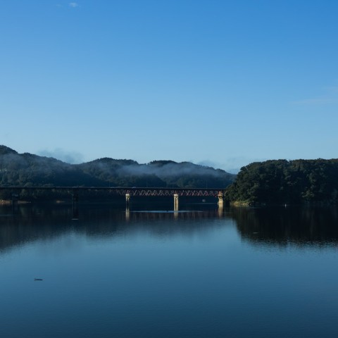 釜房ダムに釜房大橋が架かり、その姿が水面に映り込んでいます。橋の向こうには蔵王の山々がそびえています。空は澄んだ青色をしています。